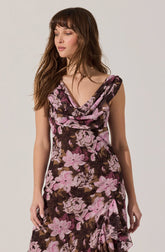 A woman is wearing a sleeveless, knee-length dress with a floral pattern. The dress has a cowl neckline and features large pink and lavender flowers with dark green and brown leaves on a dark background. The fabric appears to be soft and flowy.